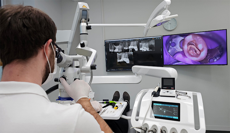 Zahnarzt arbeitet mit einem Dentalmikroskop, während auf Monitoren vergrößerte Aufnahmen eines Zahns und Röntgenbilder zu sehen sind